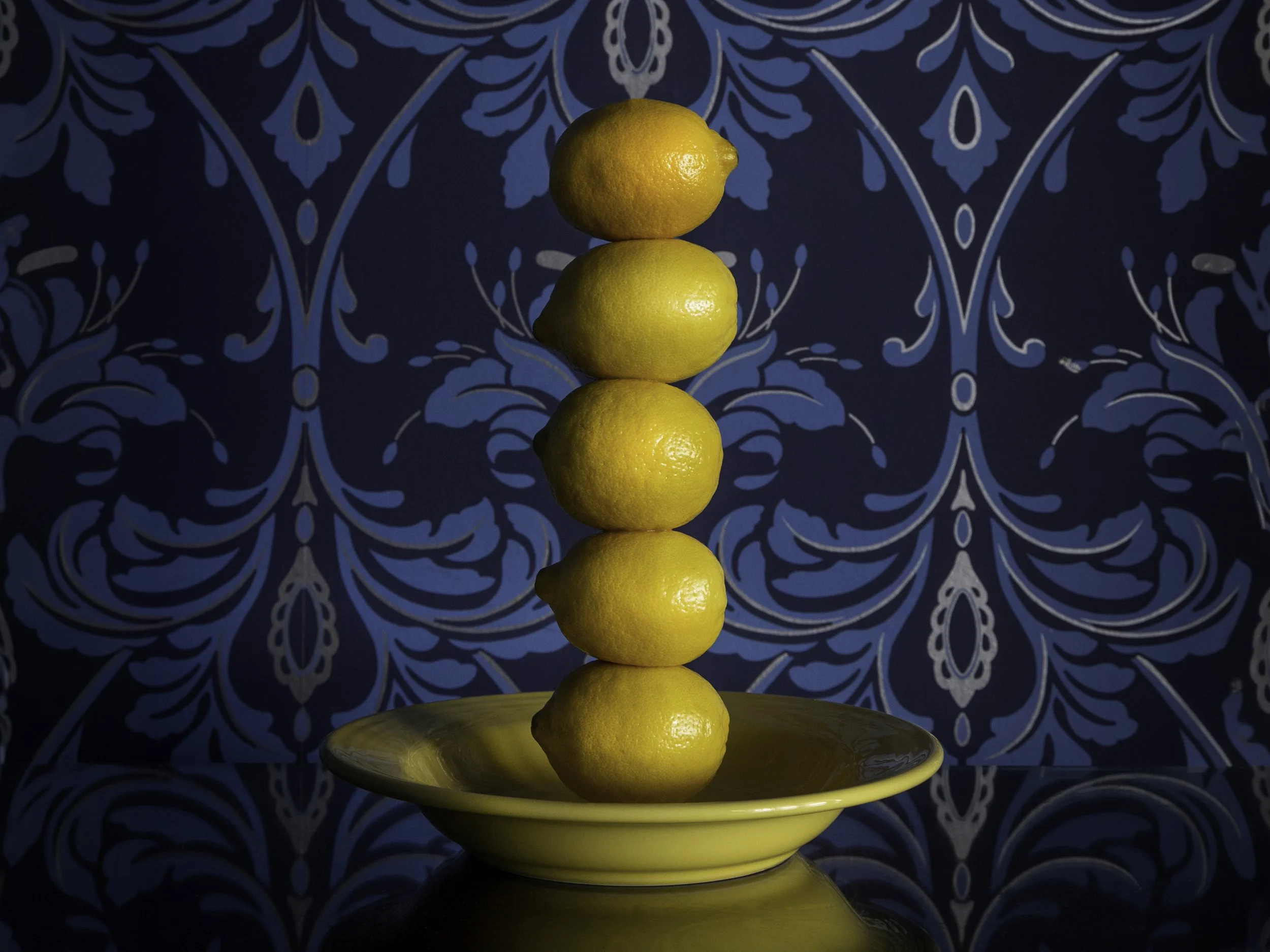 A stack of four yellow lemons arranged vertically on a yellow plate with a reflective surface. The background features a blue wallpaper with an intricate floral pattern.