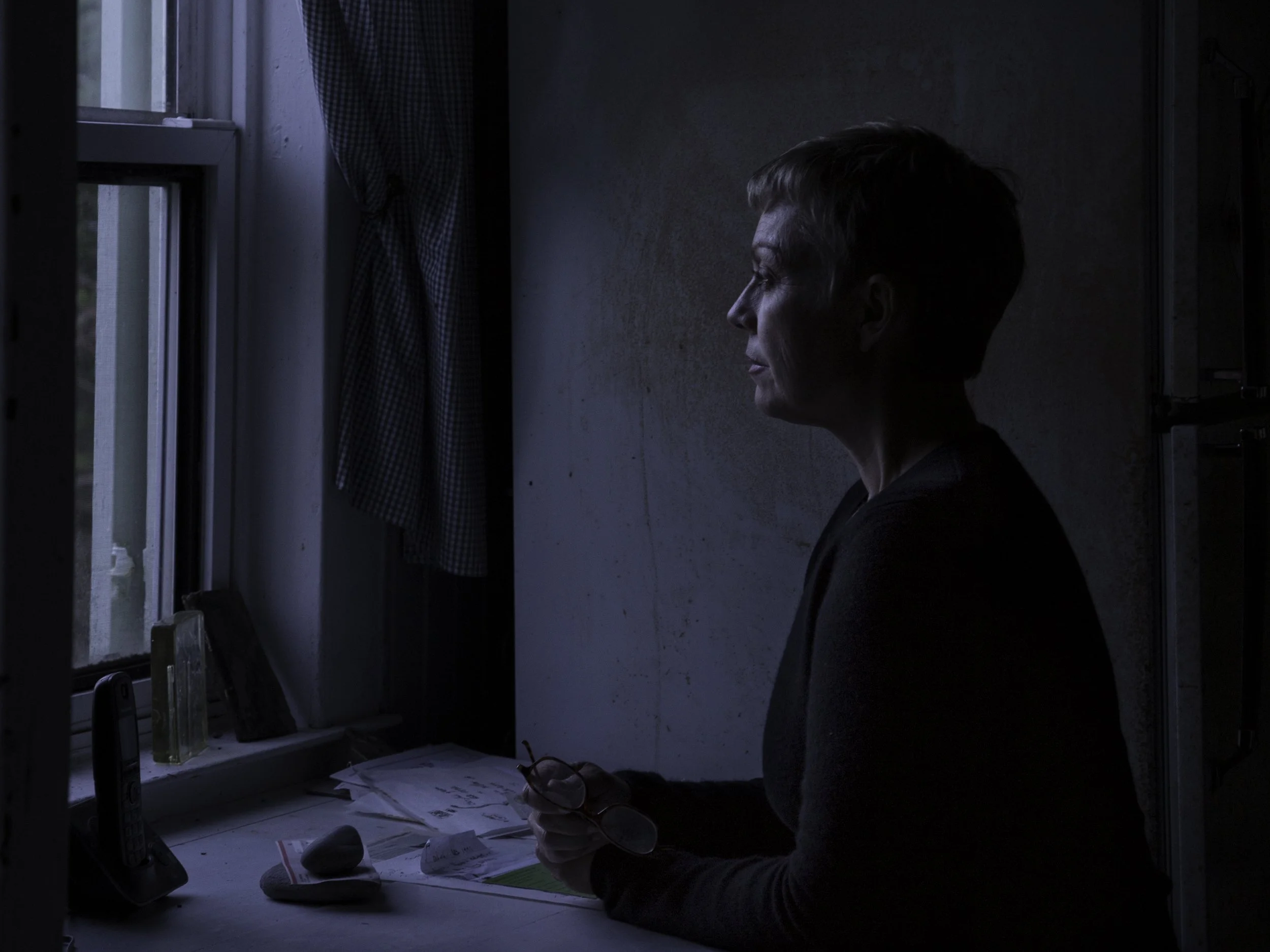 A woman with short hair sitting by a window in a dark room, writing on paperwork on a cluttered desk with glasses in her hand, illuminated by natural light coming from the window.