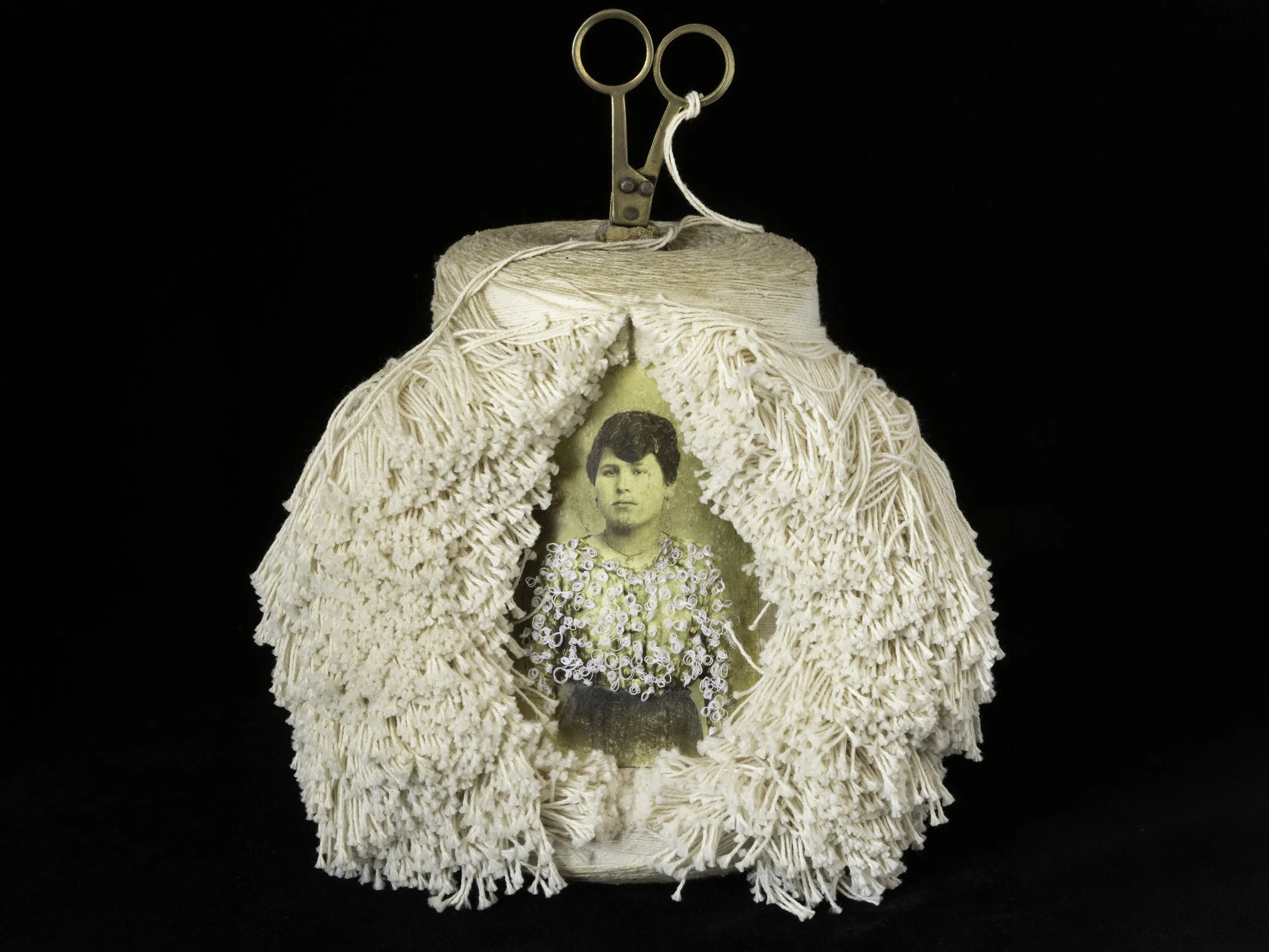 A vintage doll dressed in a cream-colored, fringed dress with a printed image of a woman on the front, set against a black background.