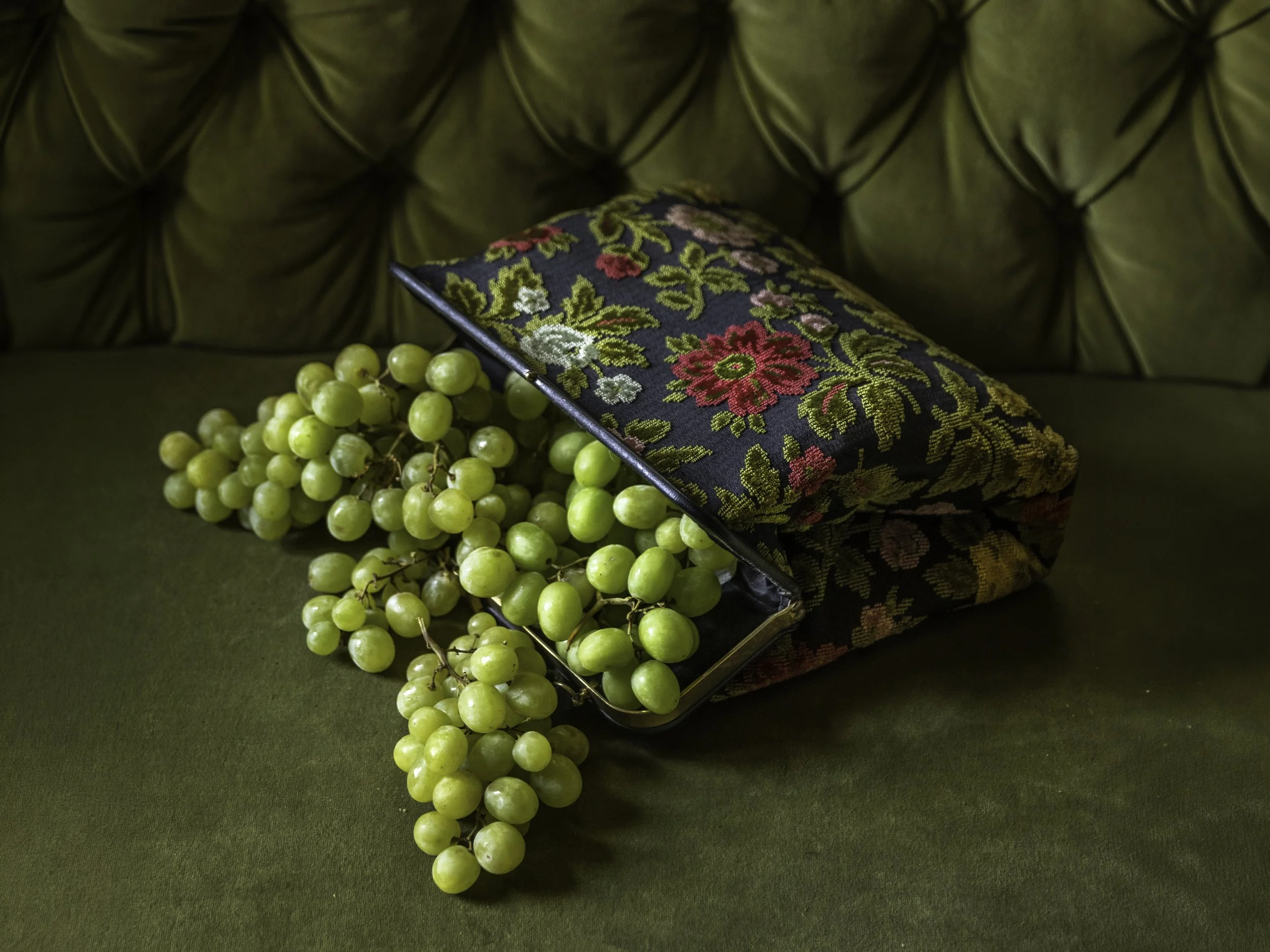 Green grapes on a dark surface with a decorative embroidered cloth and a fabric cushion with floral pattern.