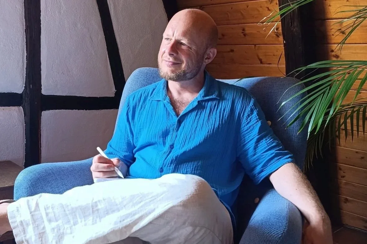 Dr. Sebastian Wolf sitzt in einem blauen Sessel in einem hellen Raum mit Fachwerk und Pflanzen.