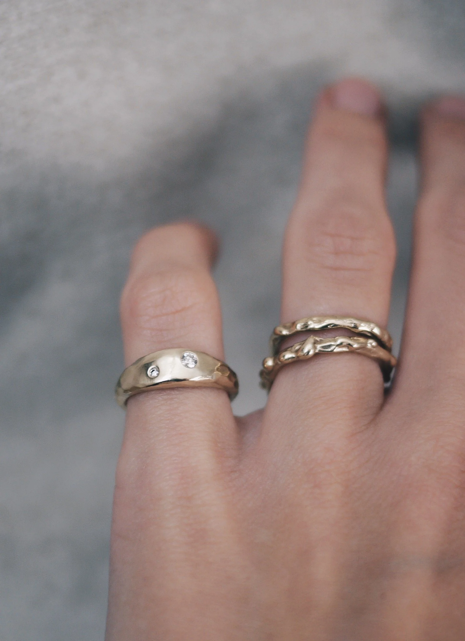 A person's hand wearing three gold and diamond rings with different designs on the middle and ring fingers, with a blurred gray background.