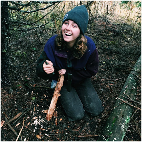 Lydia working on a feather stick, trying to carve thin curls of wood from the log that will help a fire catch