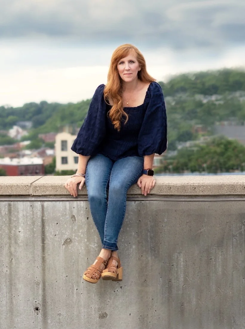 Red-haired woman sitting on a concrete ledge outdoors, wearing a navy blue blouse, blue jeans, and tan wedge sandals, with a blurred cityscape and green hills in the background.