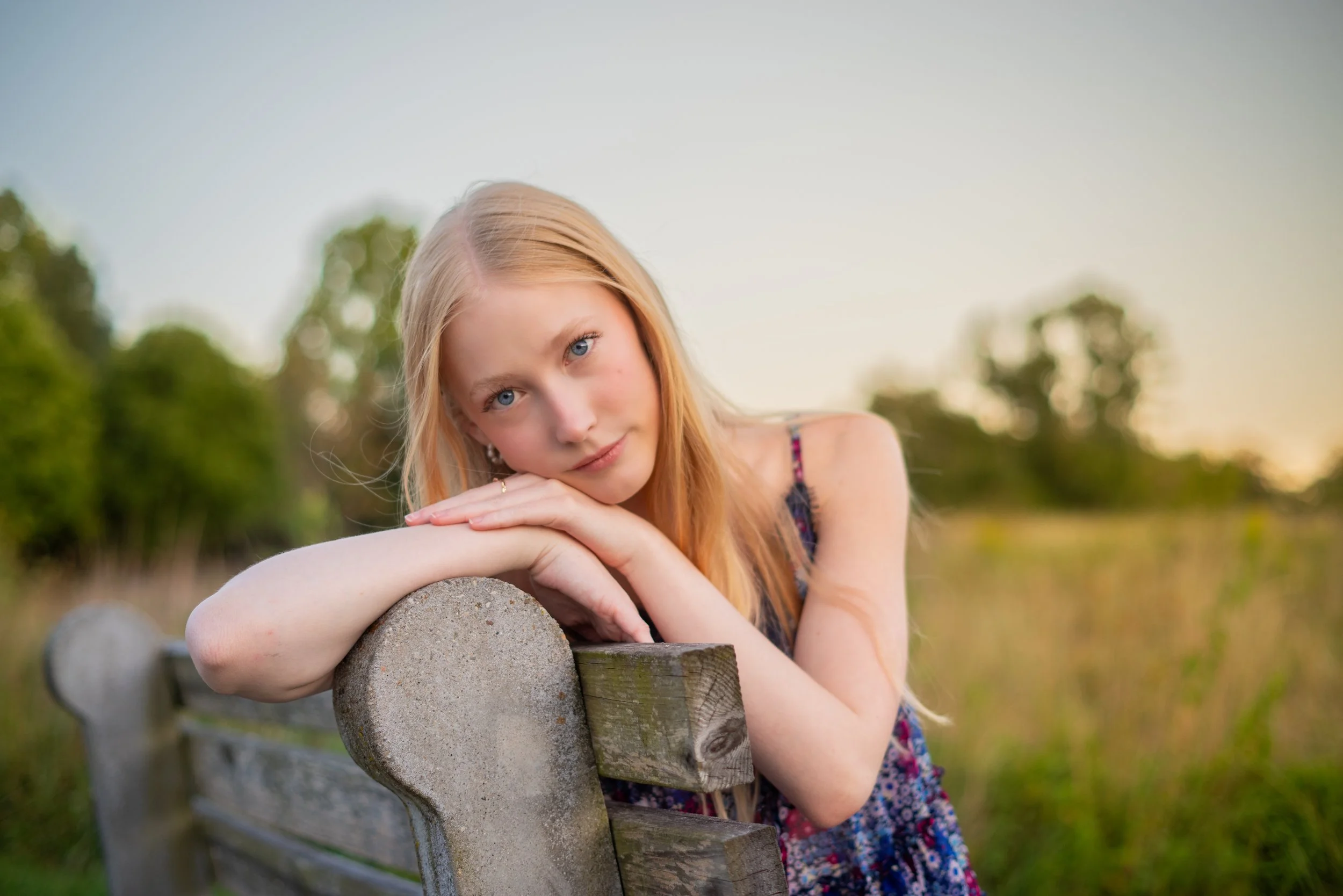 A young girl with long red hair and blue eyes resting her head on her arms on a stone and wooden park bench outdoors at sunset with trees in the background.