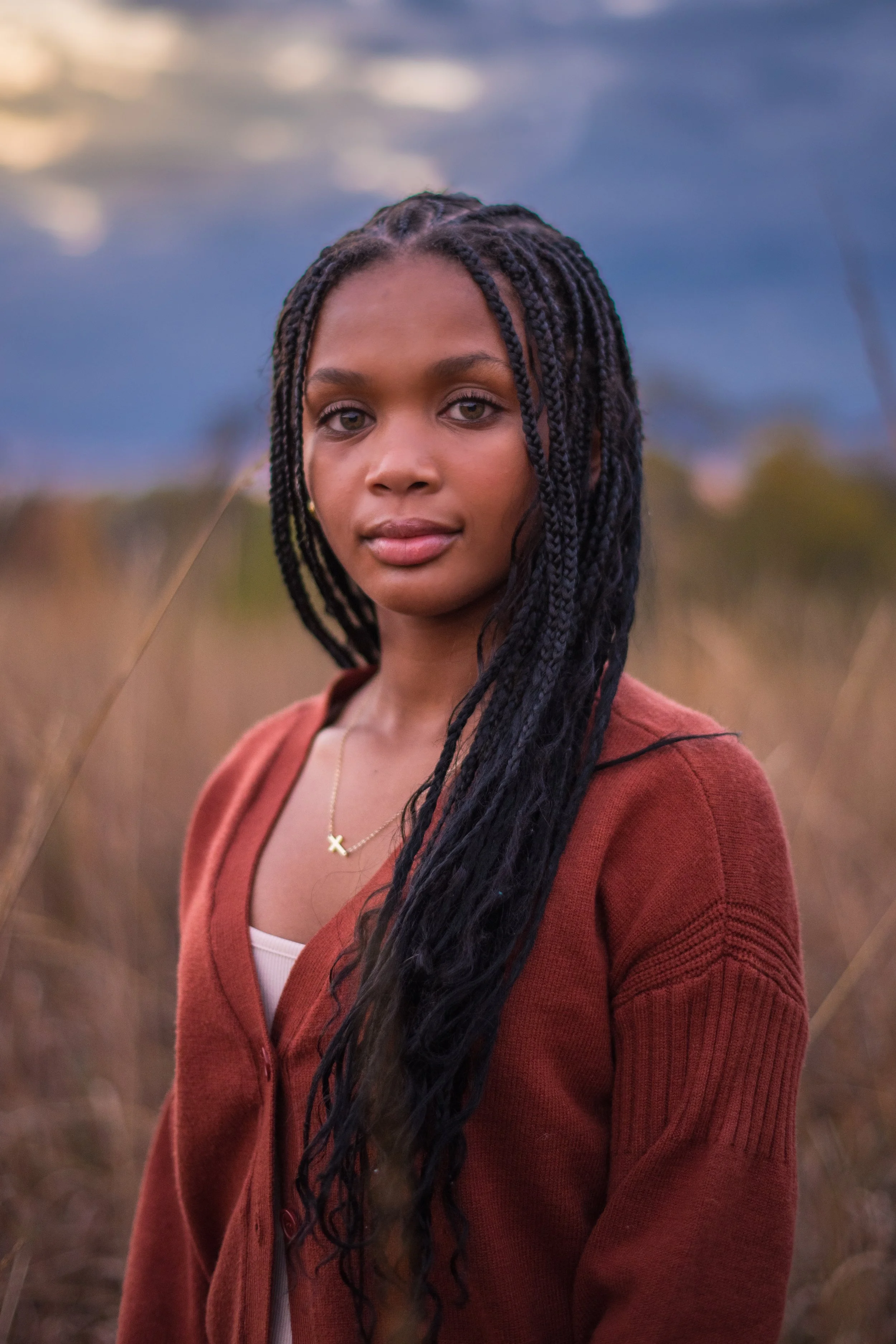 A young woman with long braided hair standing outdoors in a field during dusk, wearing a rust-colored cardigan over a white top, with a necklace featuring a cross pendant.