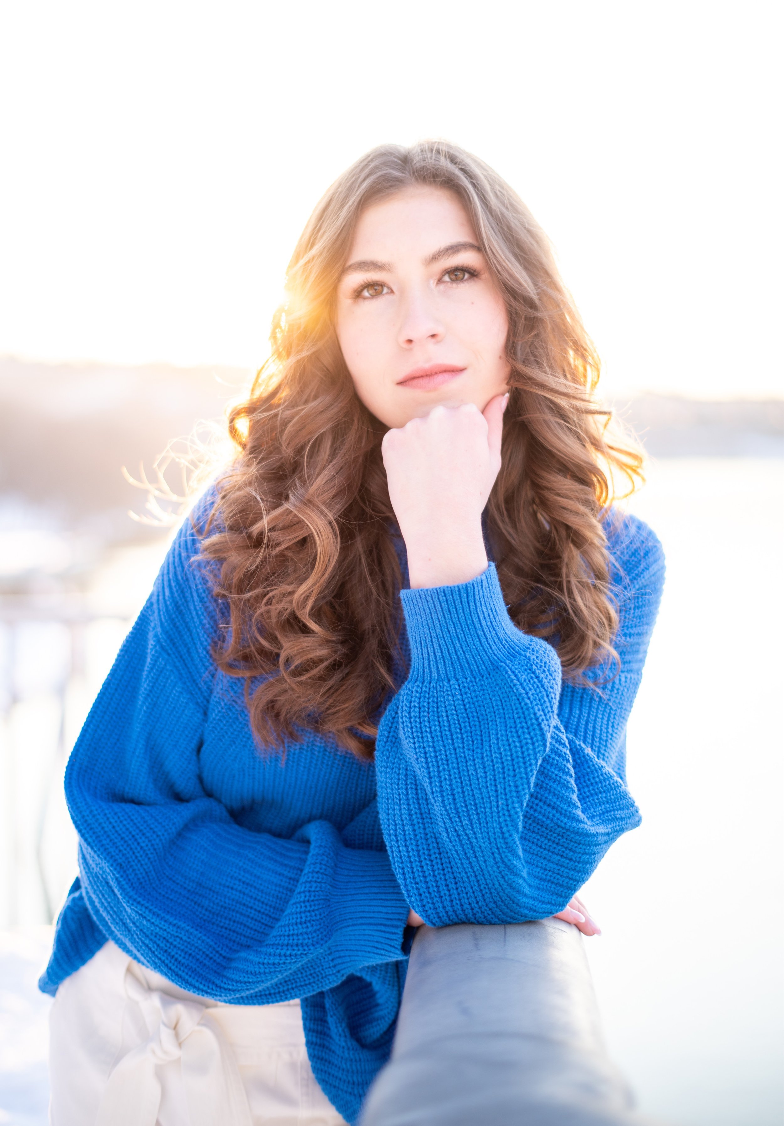 Young woman in a blue sweater resting her chin on her hand outdoors during sunset.