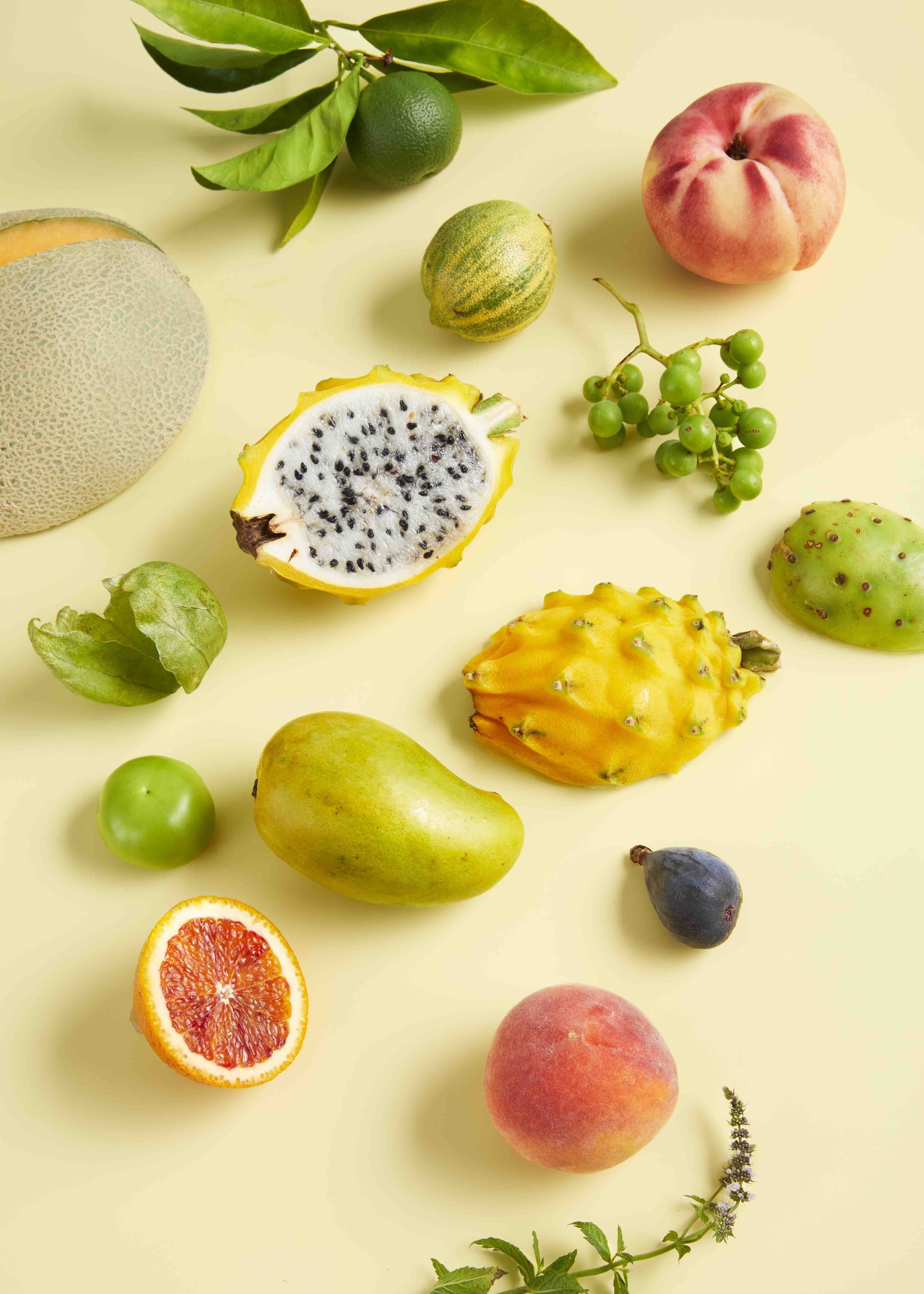 overhead shot of cut fruits, green grapes, lime with leaves, flowering herbs on yellow background.jpg