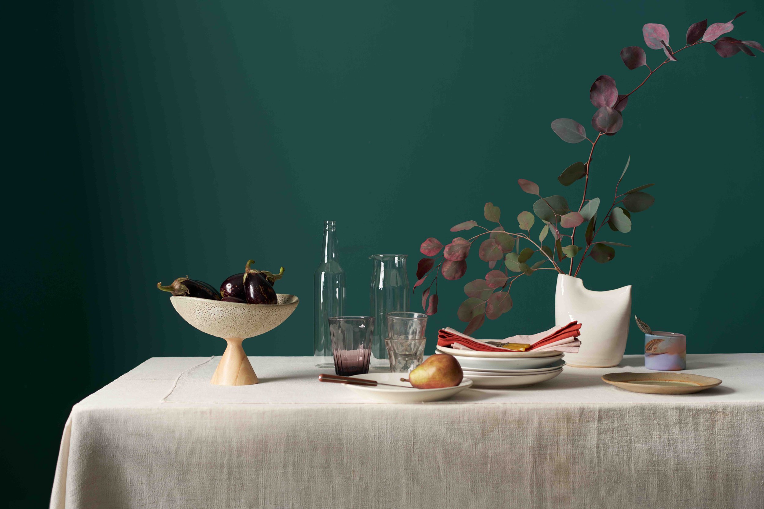 graceful organic tablescape with eggplant on a ceramic pedestal, pear and eucalyptus in front of teal wall.jpg