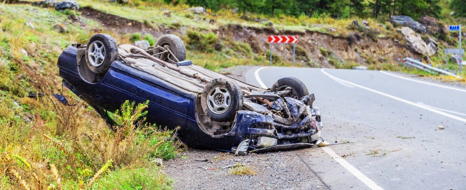 An overturned car rests on the side of a winding mountain road with safety signs and guardrails visible.
