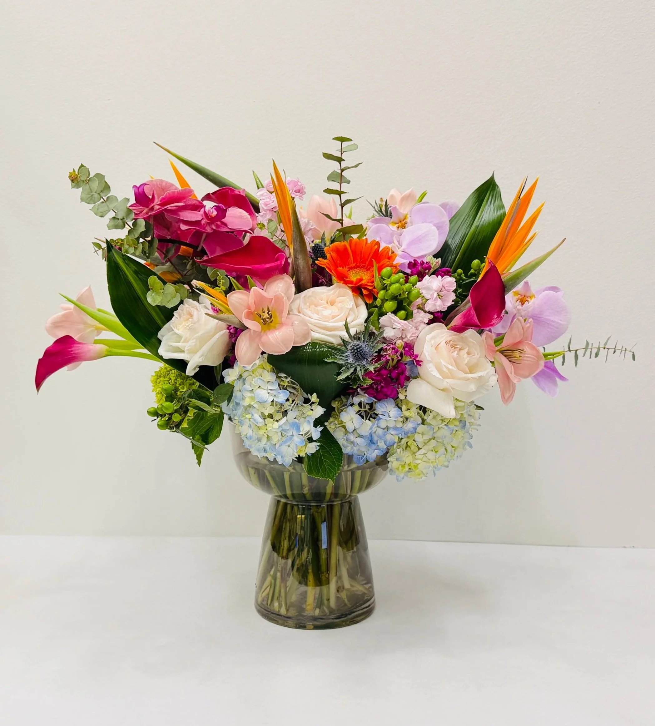 Vibrant tropical floral arrangement with roses, orchids, calla lilies, hydrangea, and birds of paradise in a modern glass vase