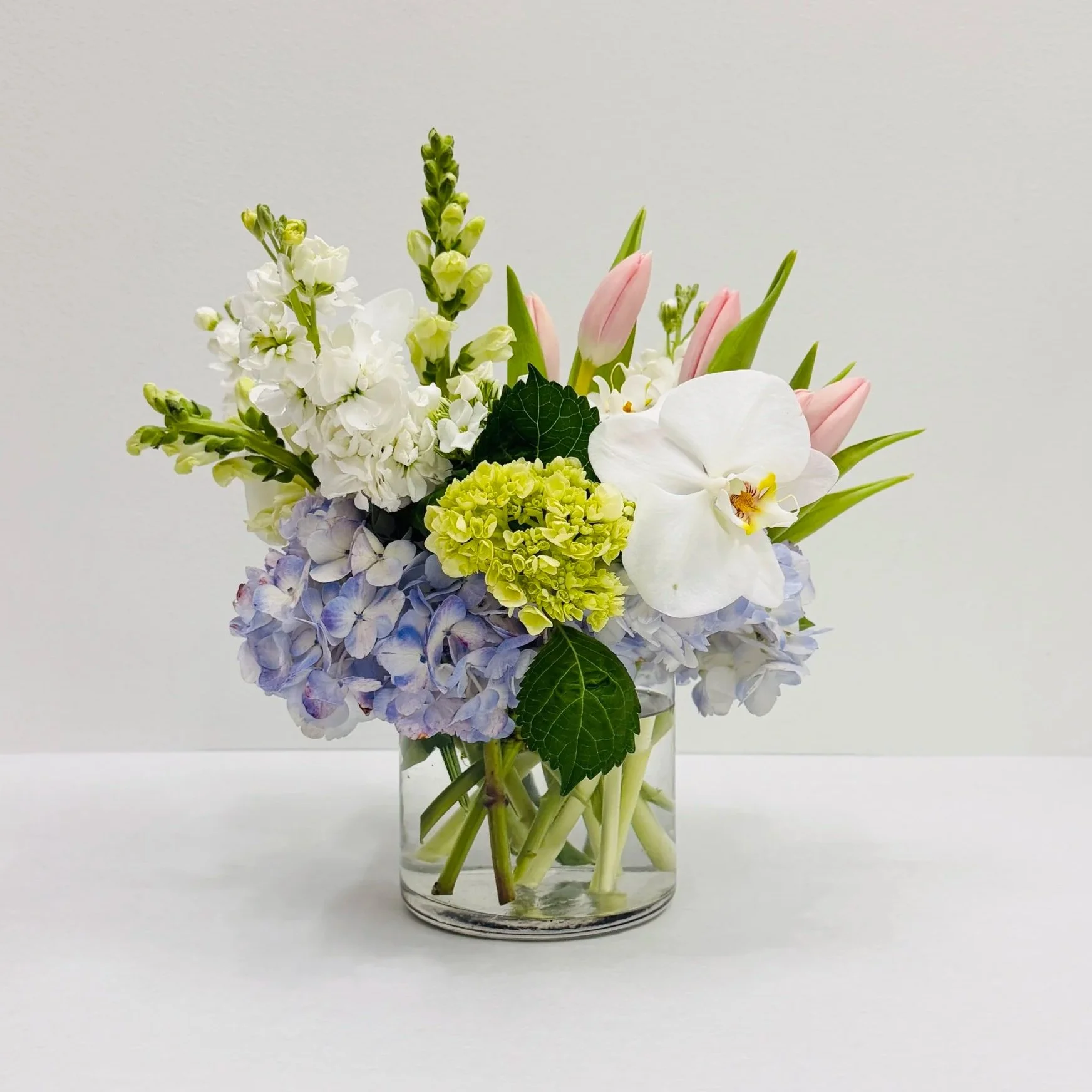 Spring pastel floral arrangement with blue hydrangea, pink tulips, white orchids, stock, and seasonal blooms in a modern clear glass cylinder vase.