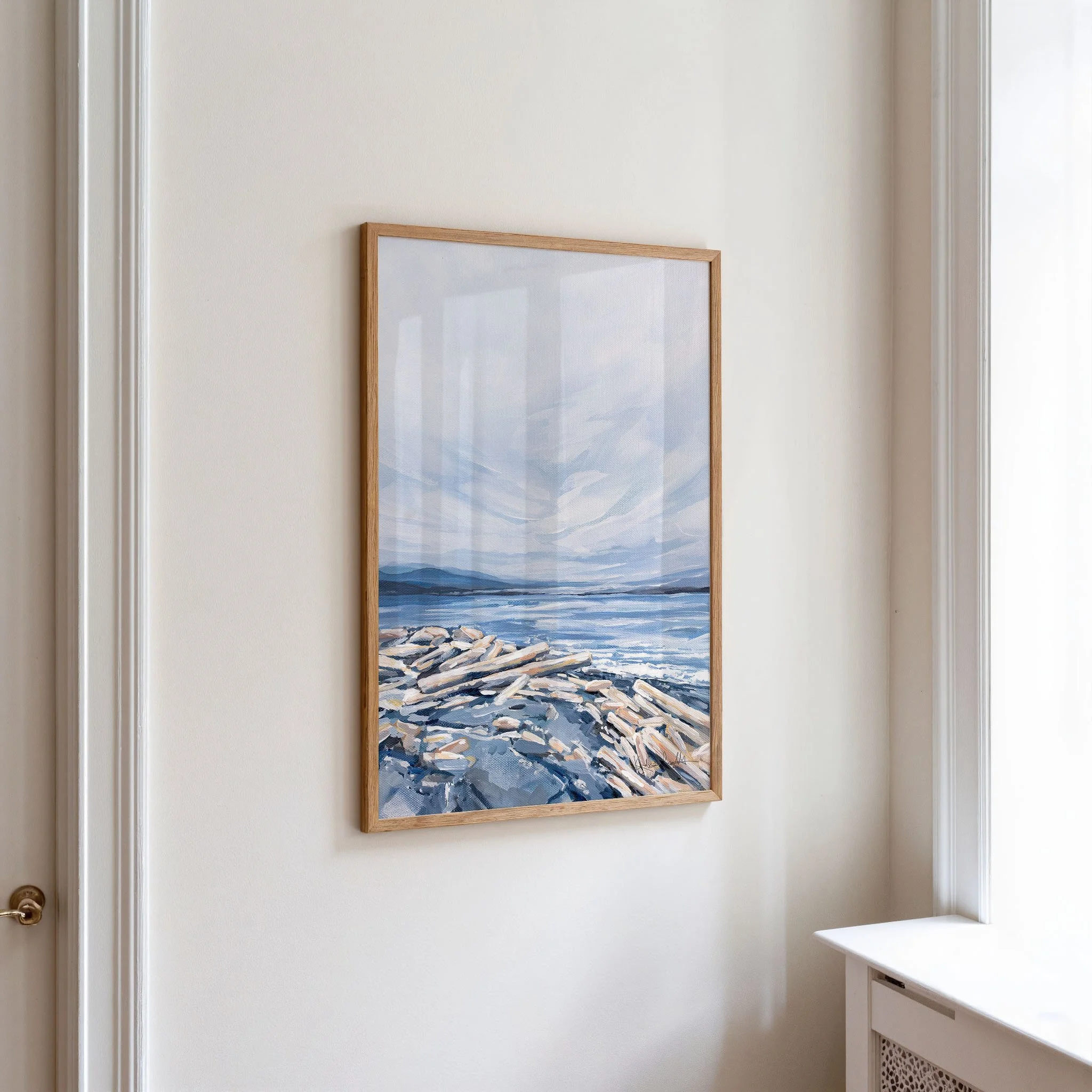 Soft coastal driftwood print in an oak frame, shown at an angle beside white peonies, calm blue seascape with distant mountains. Print by Kendra Castillo, Bellingham Washington artist.