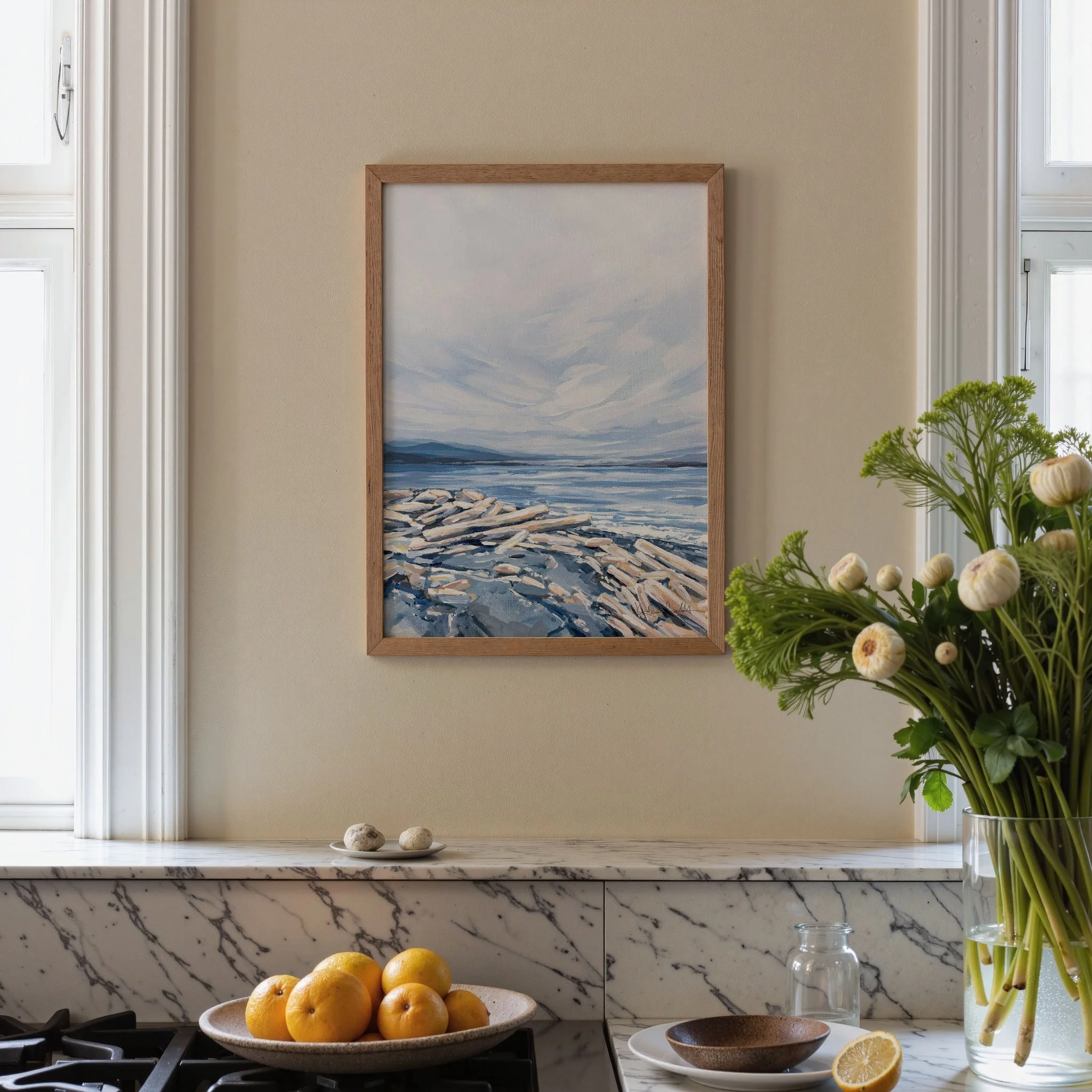 Blue and cream coastal print in an oak frame, styled above a marble kitchen counter with citrus and fresh flowers, driftwood shoreline scene. Print by Kendra Castillo, Bellingham Washington artist.