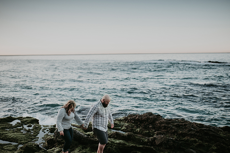 Couple walking hand in hand on mossy green rocks among the ocean.