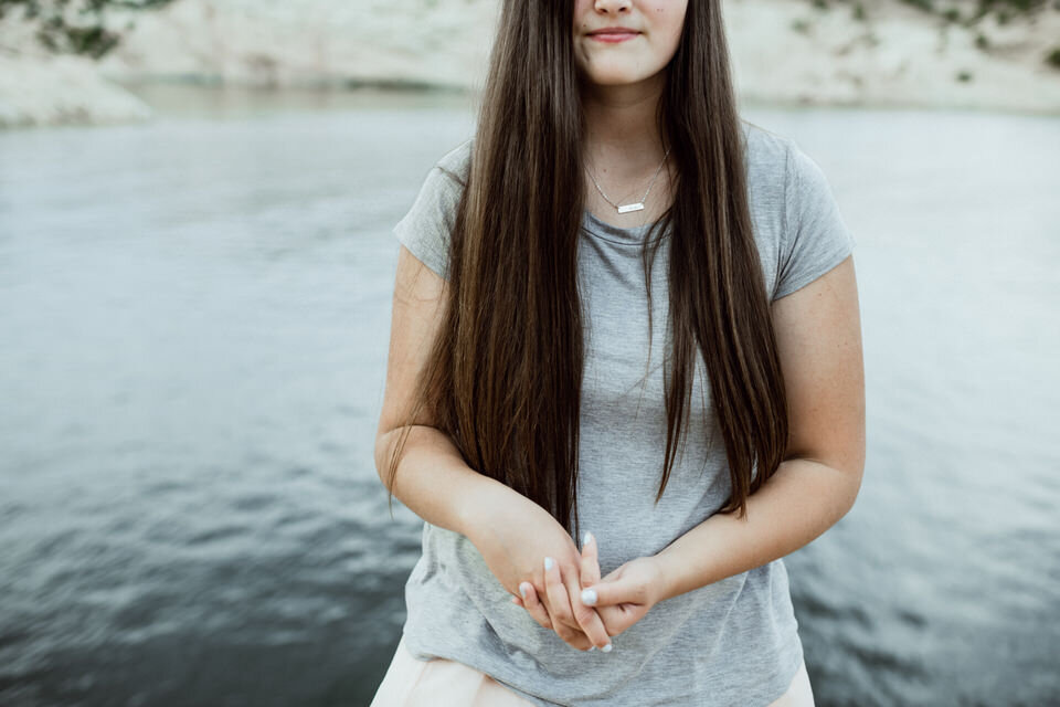 girl standing with hands clasped blue lake behind her