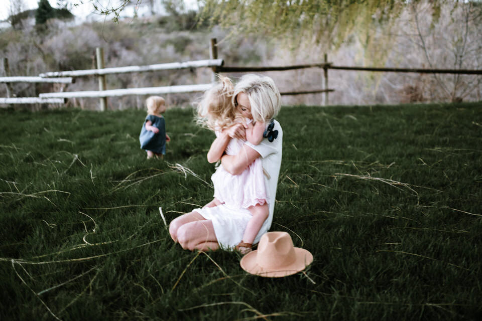 mom kneeling on grass hugging daughter while baby walks in the background