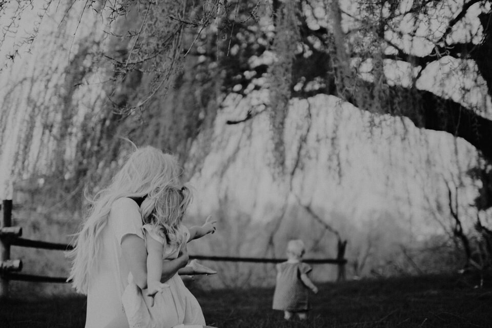 black and white photo of mom holding toddler girl looking at baby girl underneath weeping willow branches