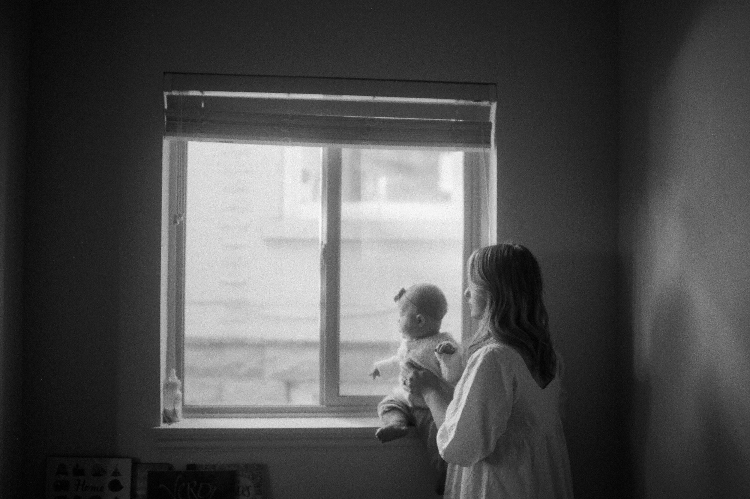 mom holding baby in windowsill looking out window