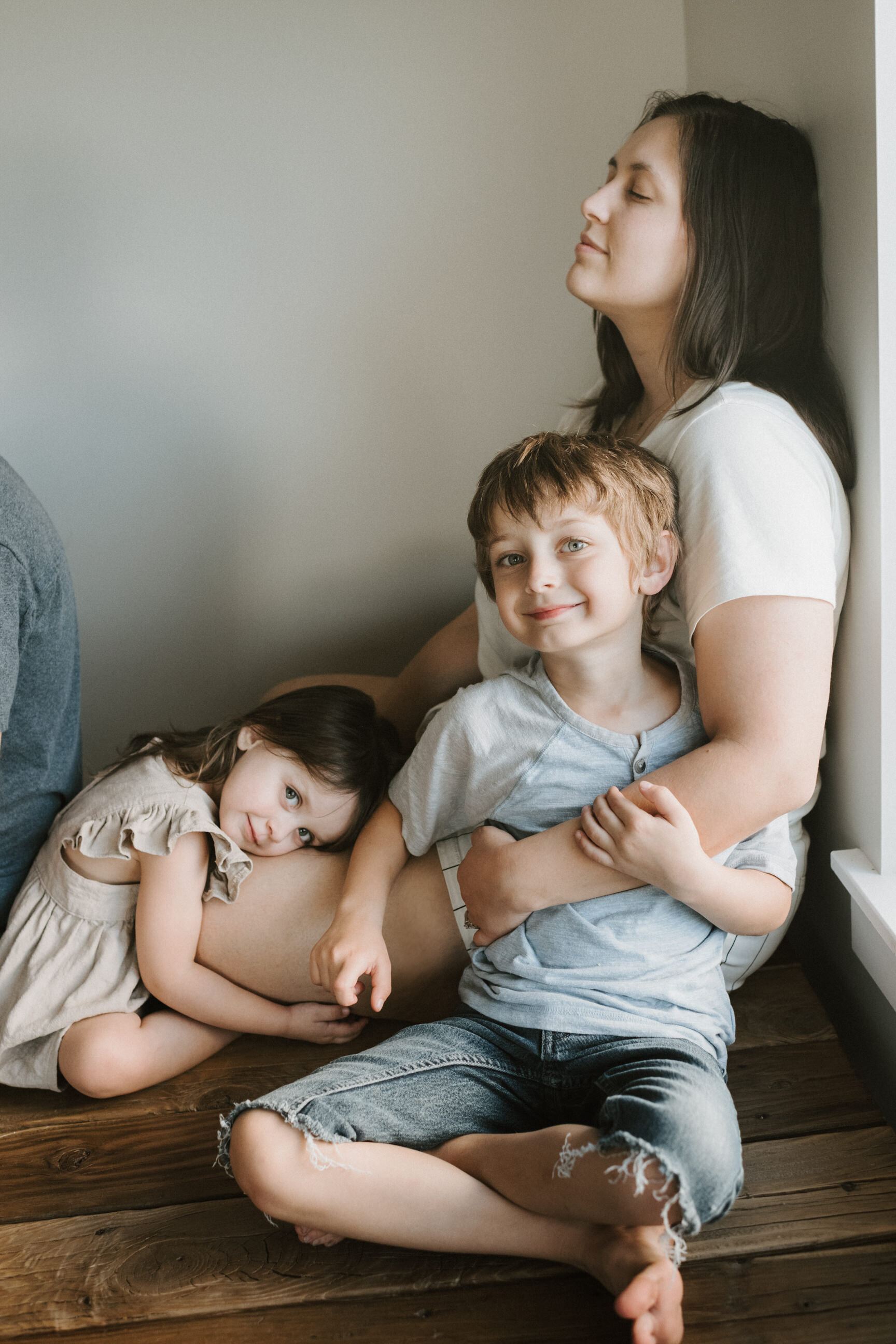 mom leaning against wall with daughter laying on her lap hugging son