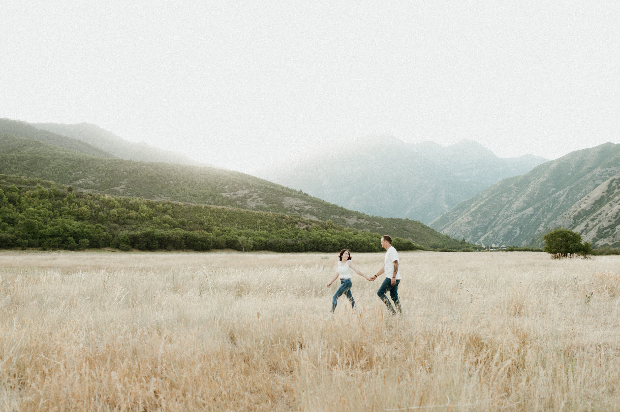 male female couple holding hands walking in golden fields with mountains in background