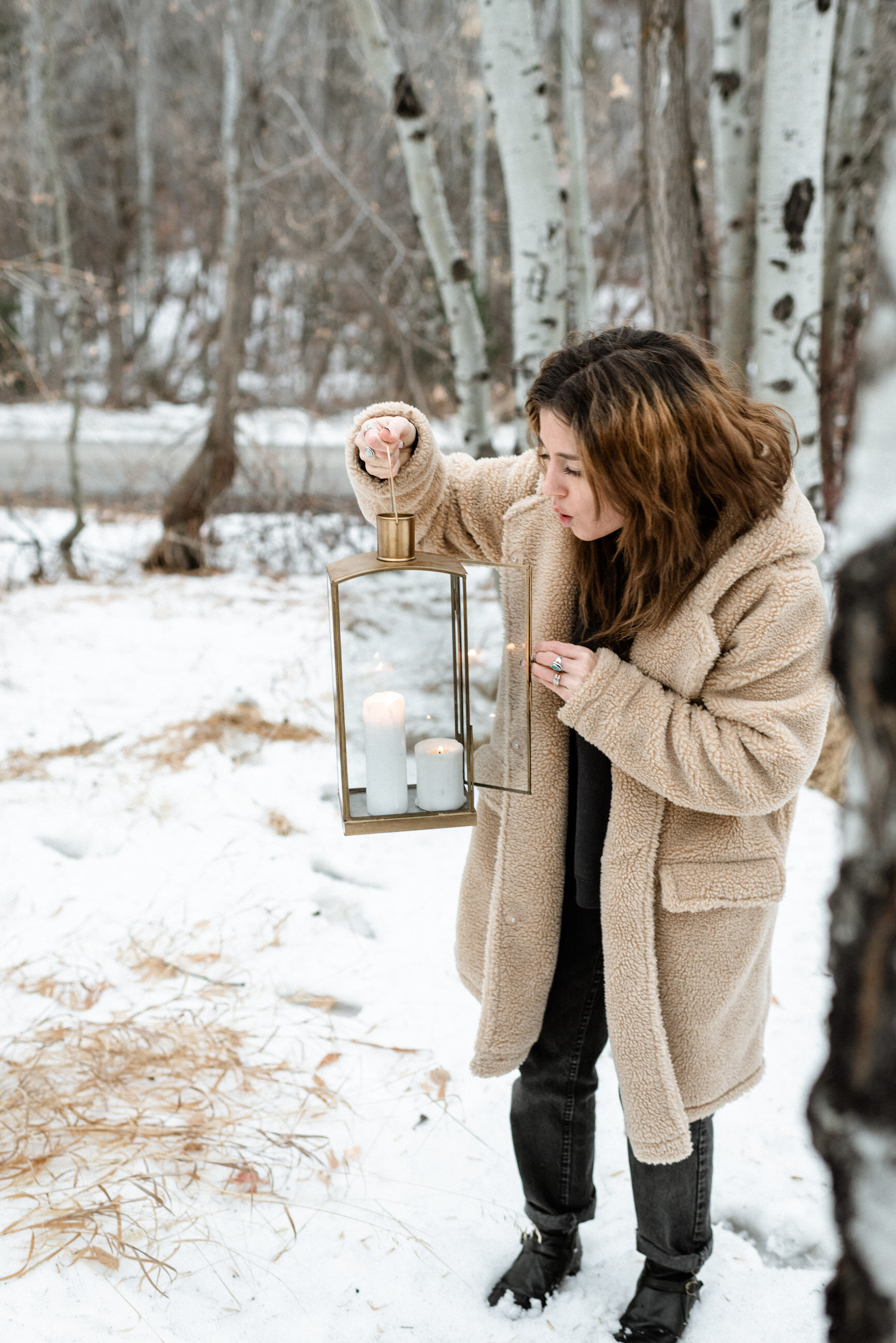 woman in fur coat holding lantern standing in snow between quaky trees