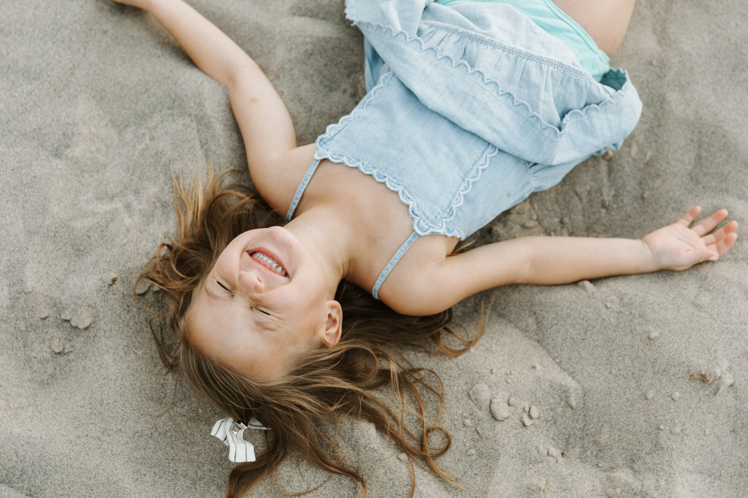 girl in blue dress lying in sand with eyes closed laughing