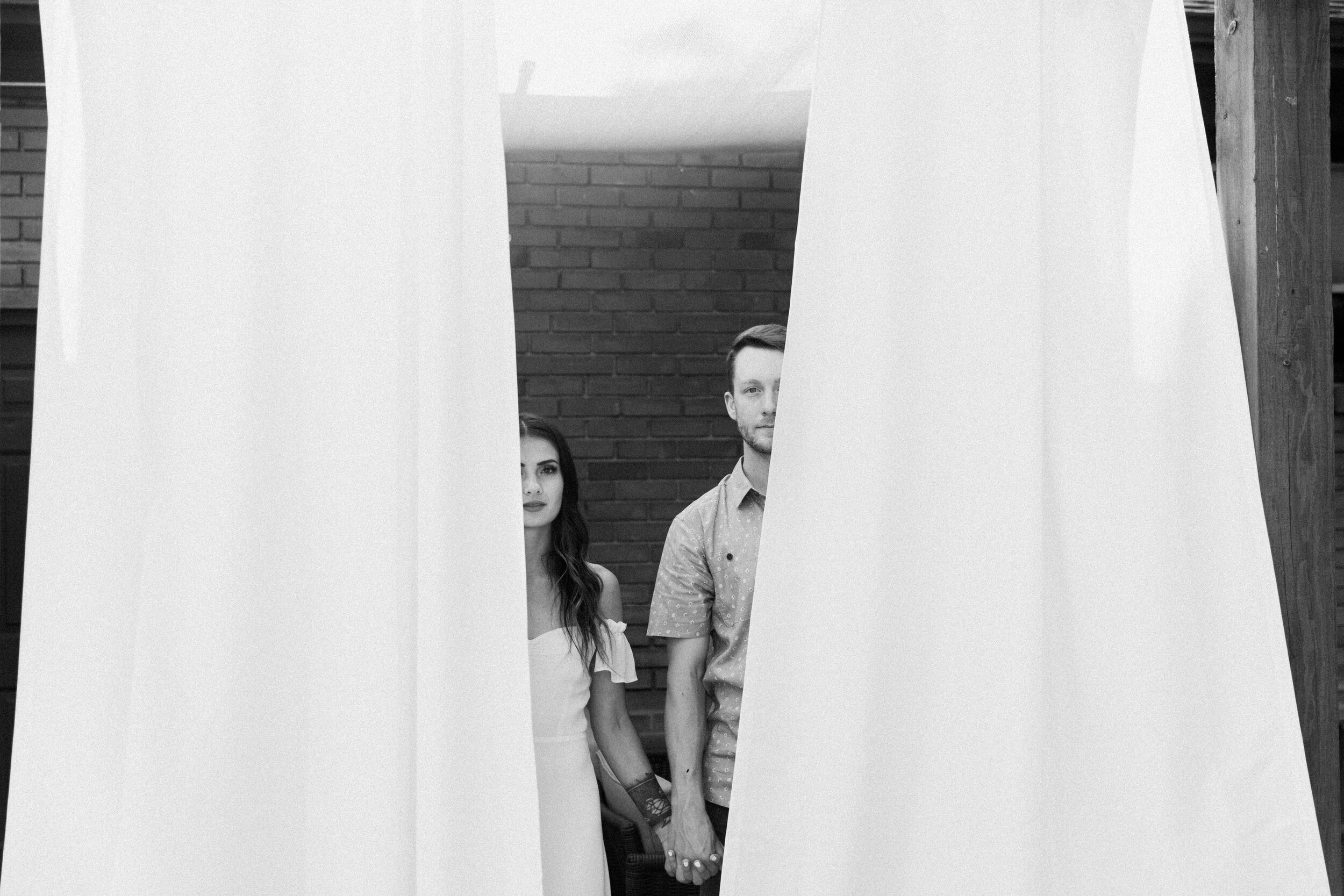 male female couple standing behind white curtains on patio