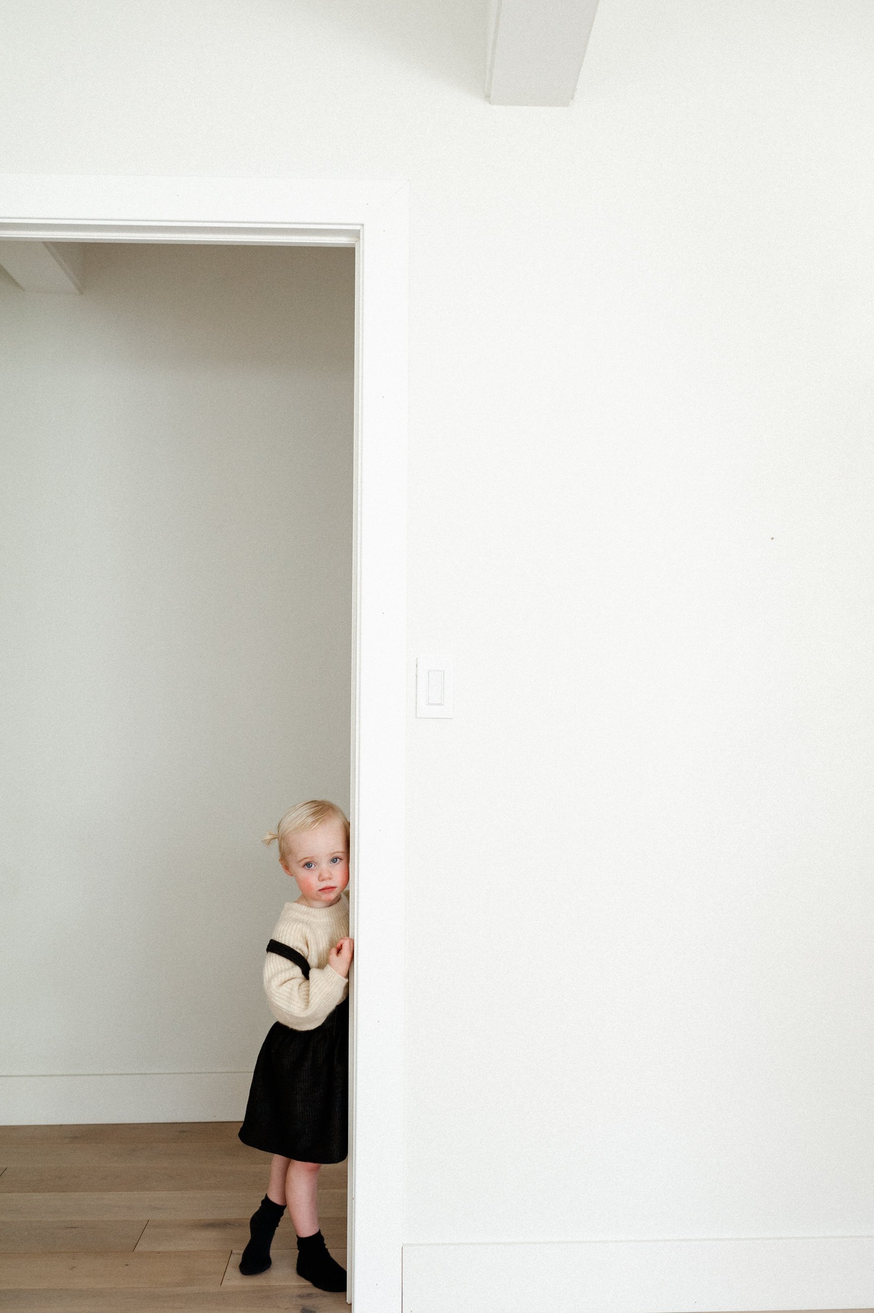 toddler girl with pigtails and overall dress leaning in white door frame