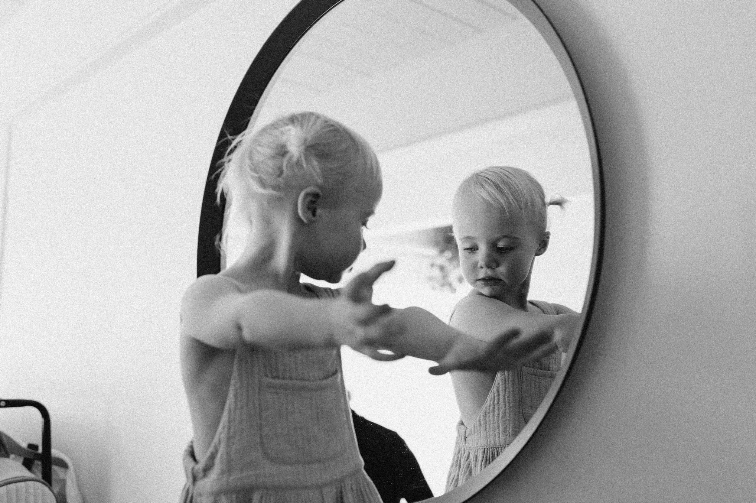 toddler girl with pig tails in overall dress looking in mirror