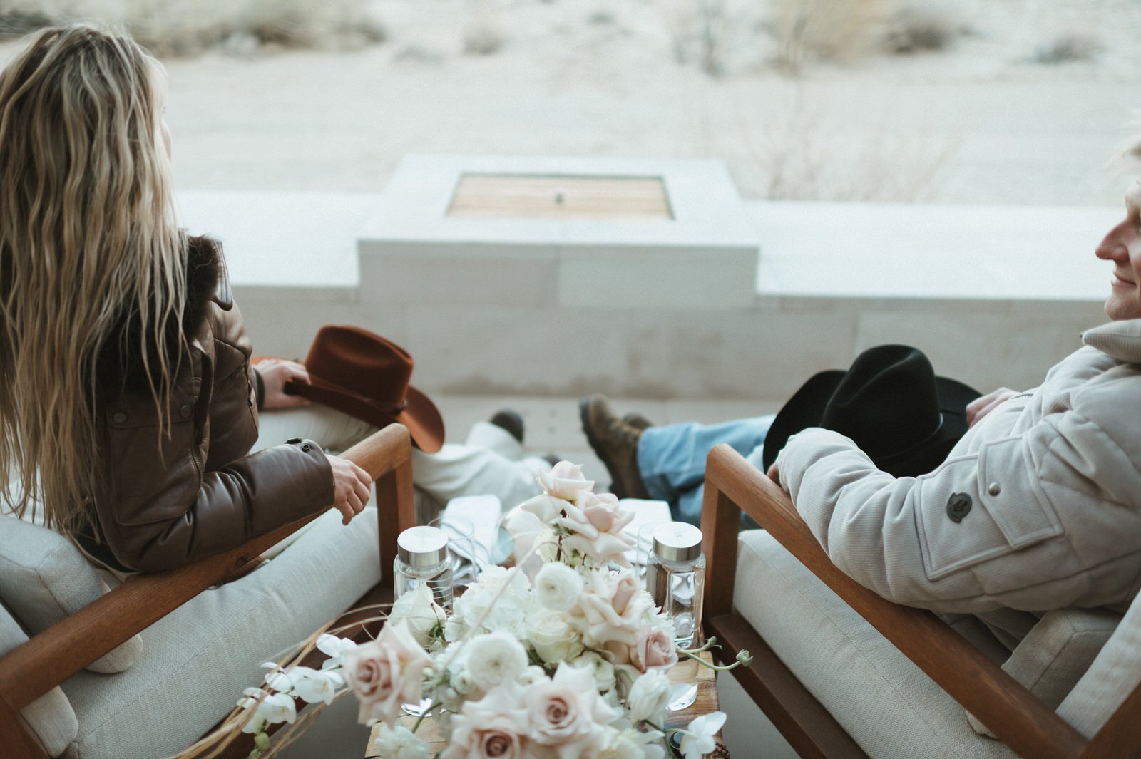 black and white film of couple sitting on veranda in white chairs wooden handles cowboy hats in laps flowers on wooden table viewing desert landscape and sunset at amangiri