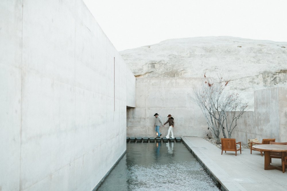 couple wearing western wear cowboy hats walking on concrete sidewalk surrounded by concrete walls in front of blue water at amangiri