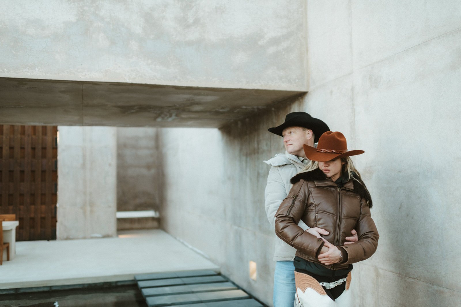 couple hugging brown leather jacket white chaps cowboy hats concrete walls at amangiri