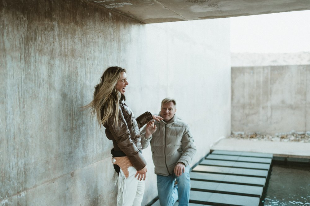man kneeling down proposing to woman laughing holding hands on blue steps concrete walls amangiri
