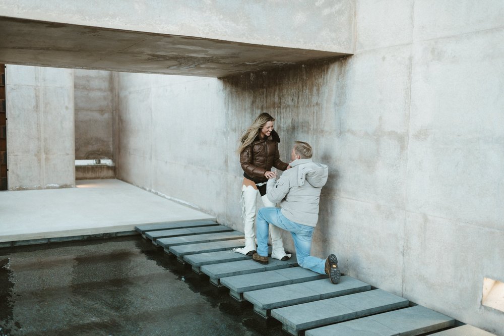 man kneeling down proposing to woman laughing holding hands on blue steps concrete walls amangiri