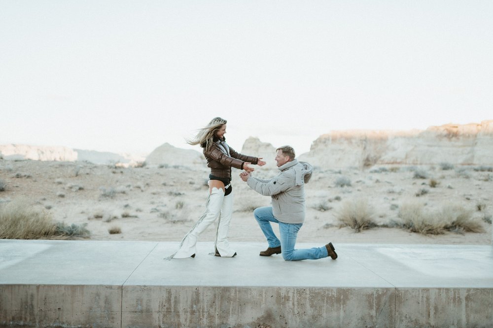 man kneeling down proposing to woman on concrete wall with desert landscape and mesas in the background amangiri