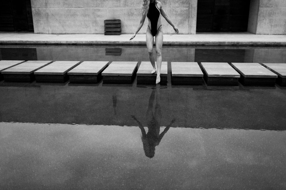 black and white photo woman in black swimsuit standing on steps of reflection pool amangiri touching toes into water
