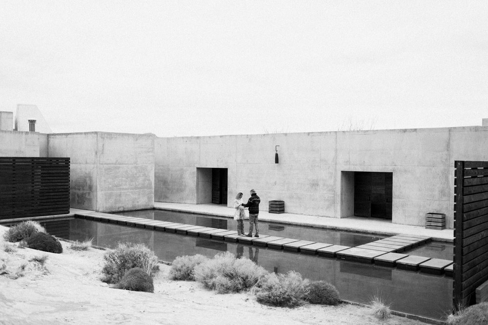 black and white photo man and woman holding hands on steps across pool concrete walls desert plants and sand