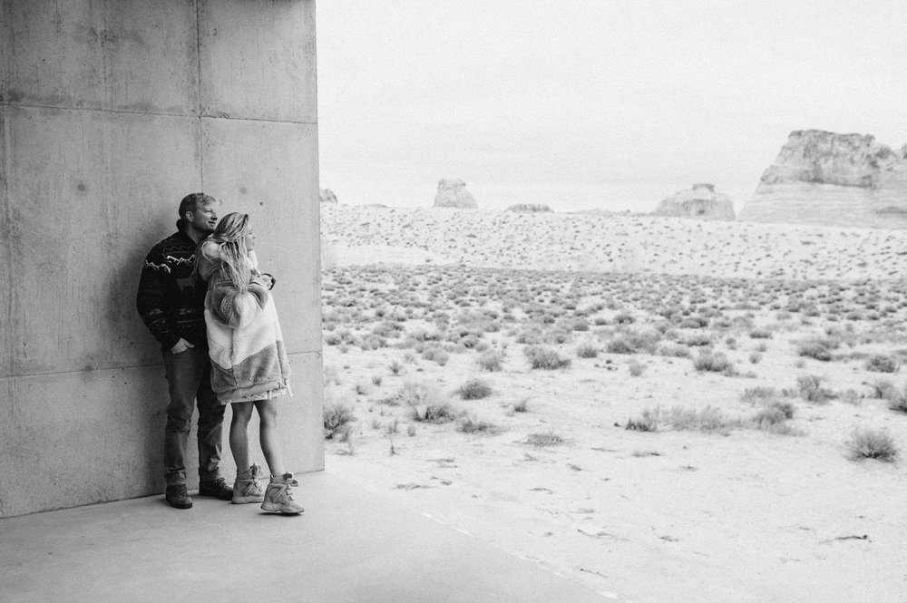 black and white photo man and woman hugging both looking toward desert standing on concrete leaning against concrete wall amangiri