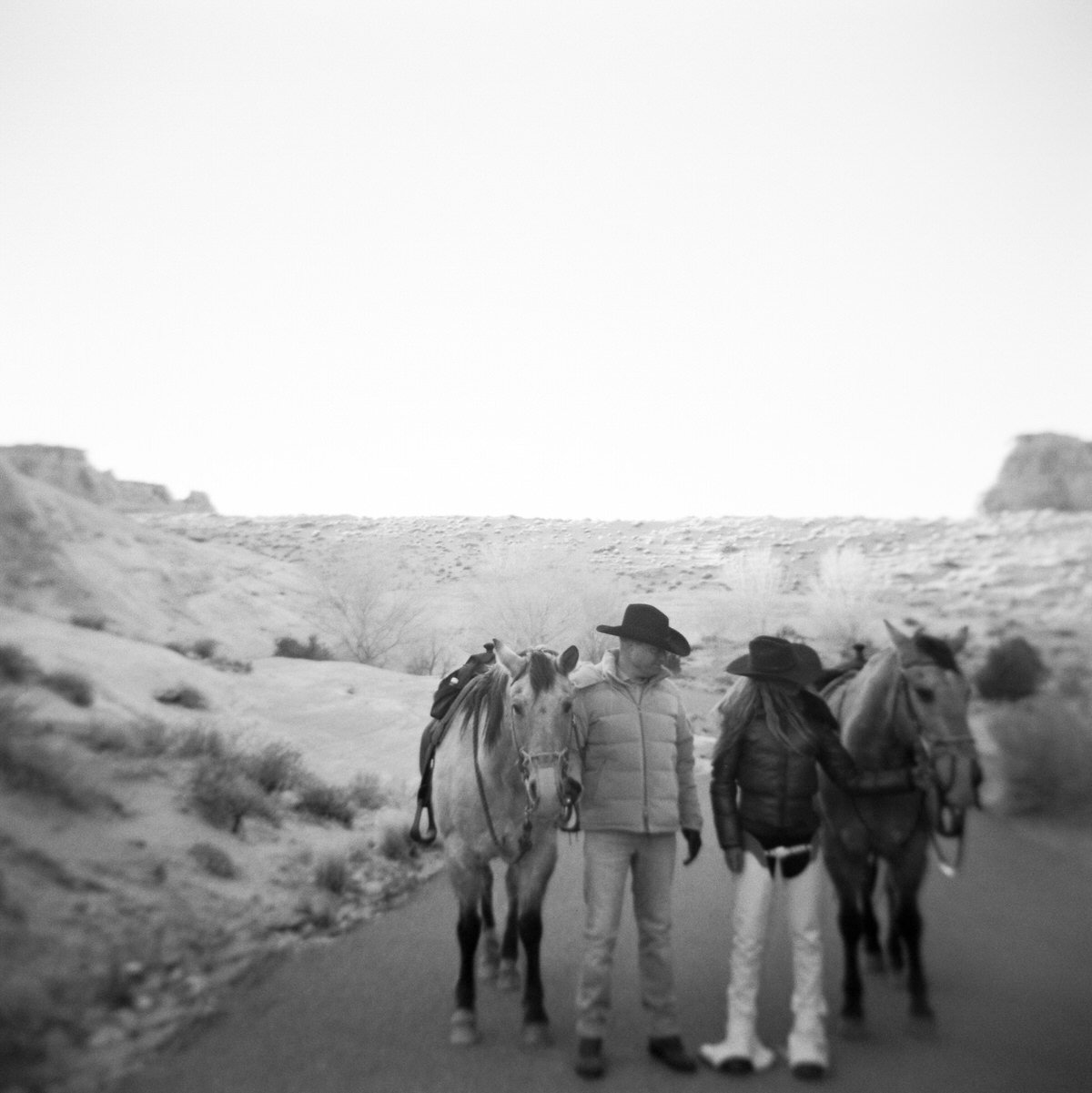 black and white film of couple standing in winding road next to horses at amangiri