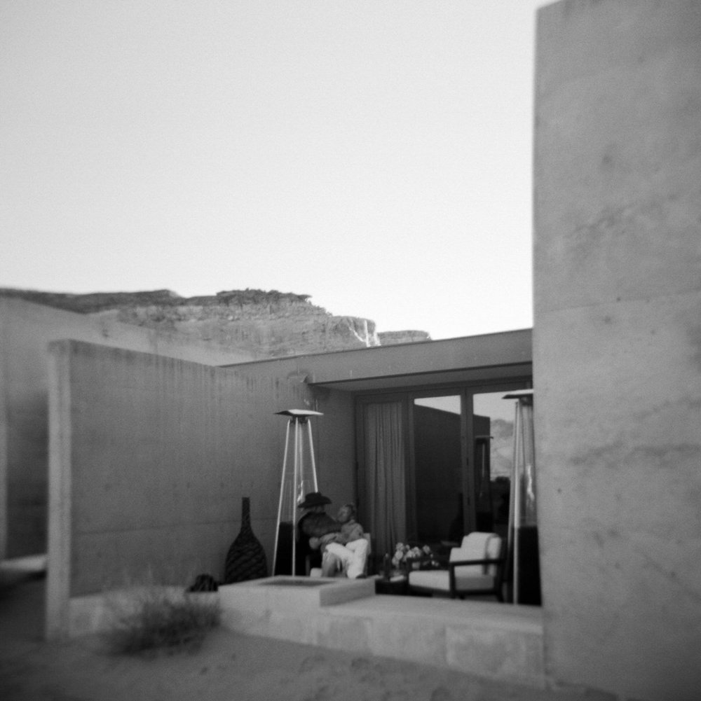 black and white film of couple sitting on veranda in white chairs wooden handles cowboy hats in laps flowers on wooden table viewing desert landscape and sunset concrete walls at amangiri resort