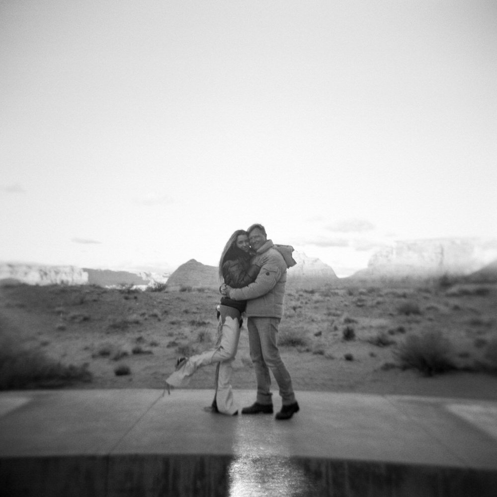 black and white square film photo man and woman hugging with desert landscape and mesas background amangiri