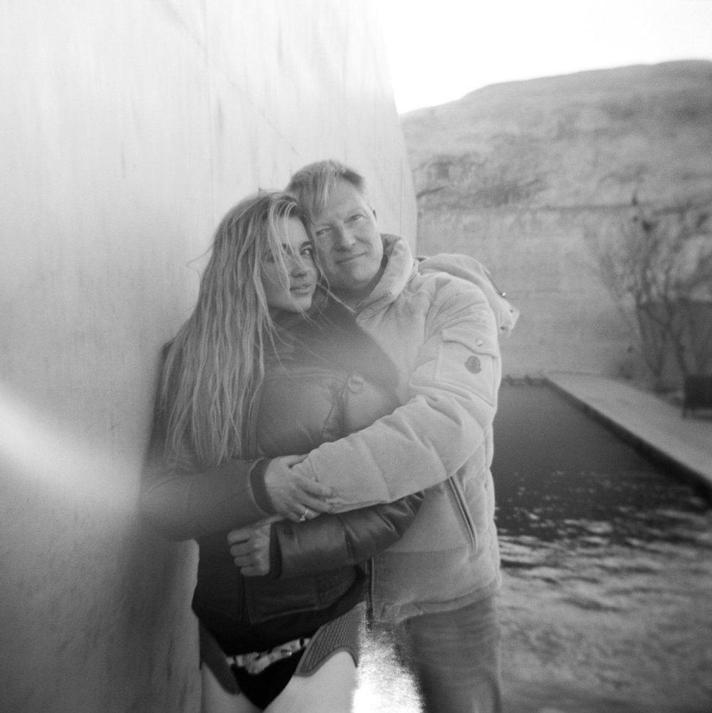 black and white film square photo couple hugging facing camera winter coats in front of concrete wall amangiri