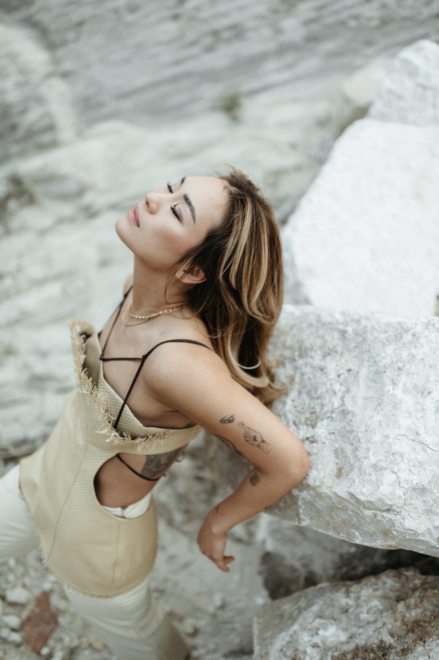 asian woman of color in straw editorial top with cutouts black ribbon leaning arms on rock in desert eyes closed chin tilted upward