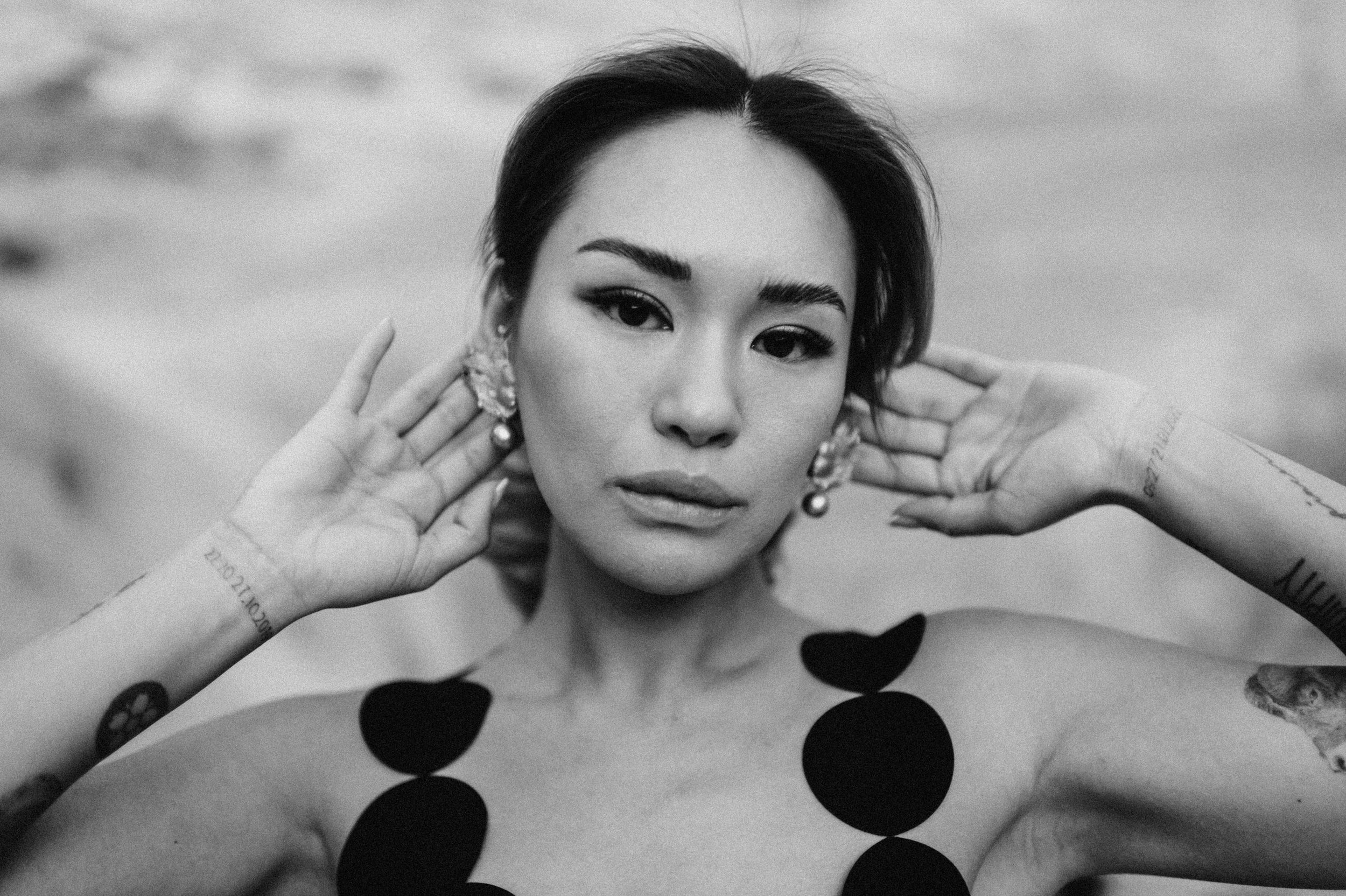 close up woman of color black bold eyeliner in black circle cutout dress hands behind ears looking at camera hands behind ears showcasing bold silver custom earrings