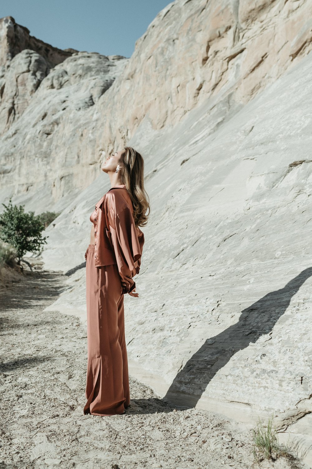 woman in desert in mauve silk loungewear hands behind back shadow on rock behind her eyes closed face tilted toward sun