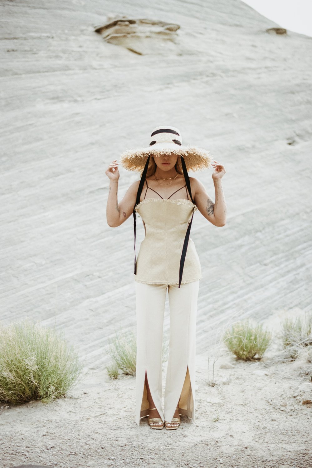 asian woman of color in straw editorial hat and top with cutouts black ribbon leaning on rock in desert
