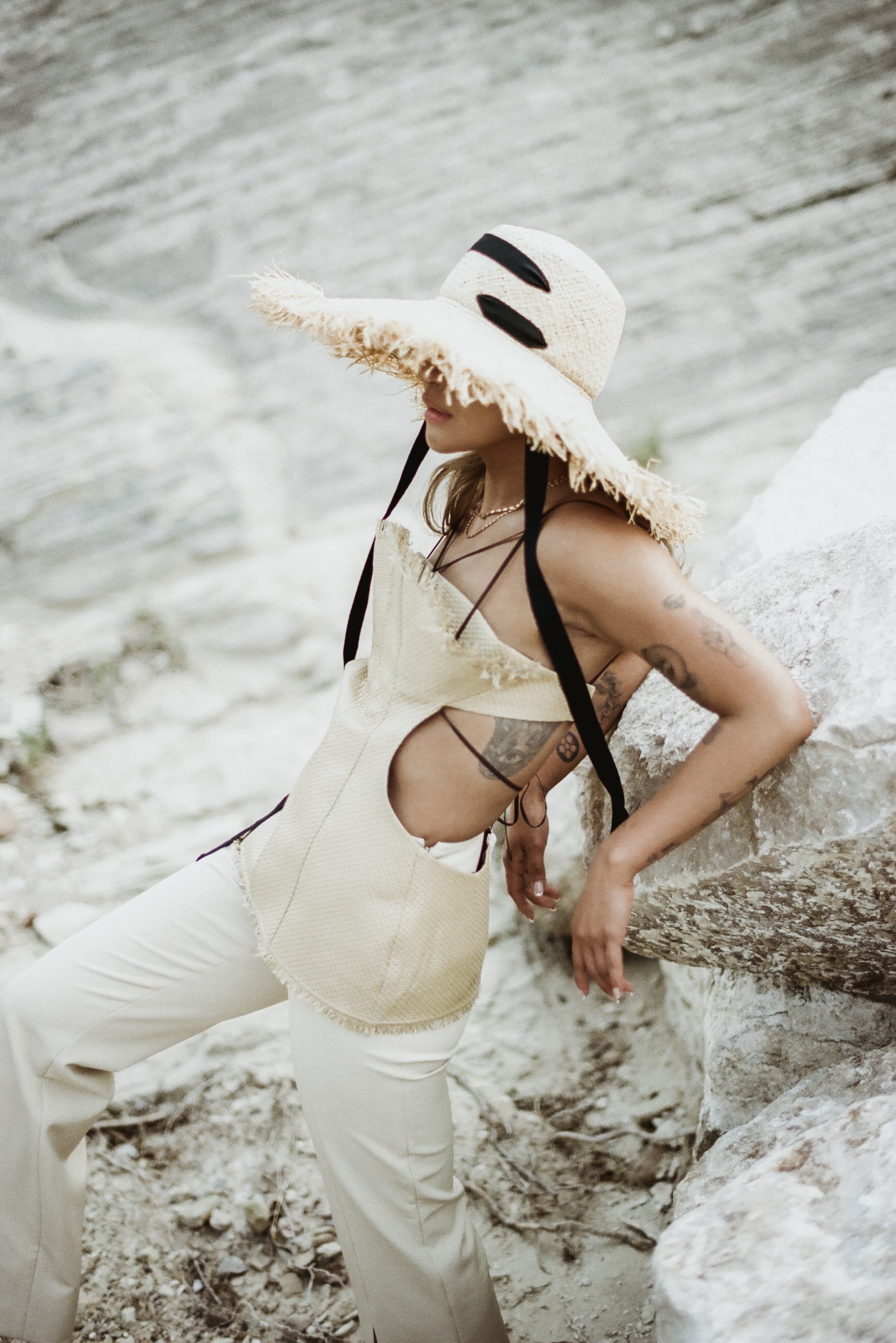 asian woman of color in straw editorial hat and top with cutouts black ribbon leaning on rock in desert