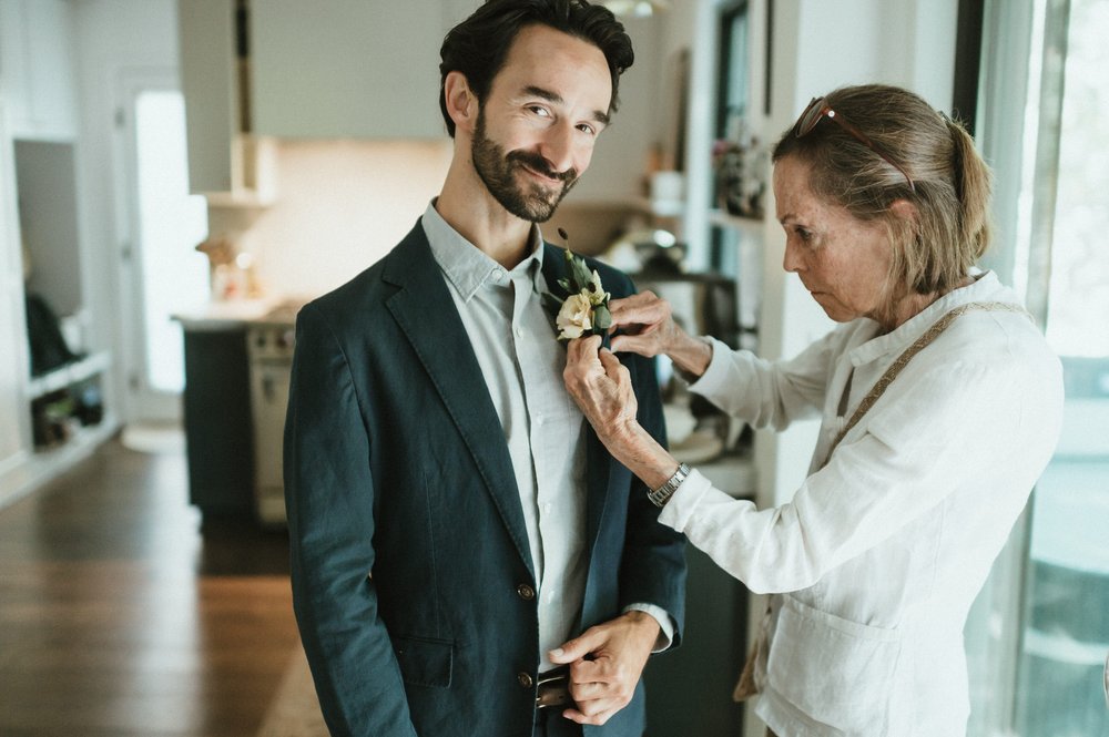groom in suit with grey shirt getting boutonniere pinned on by mother 