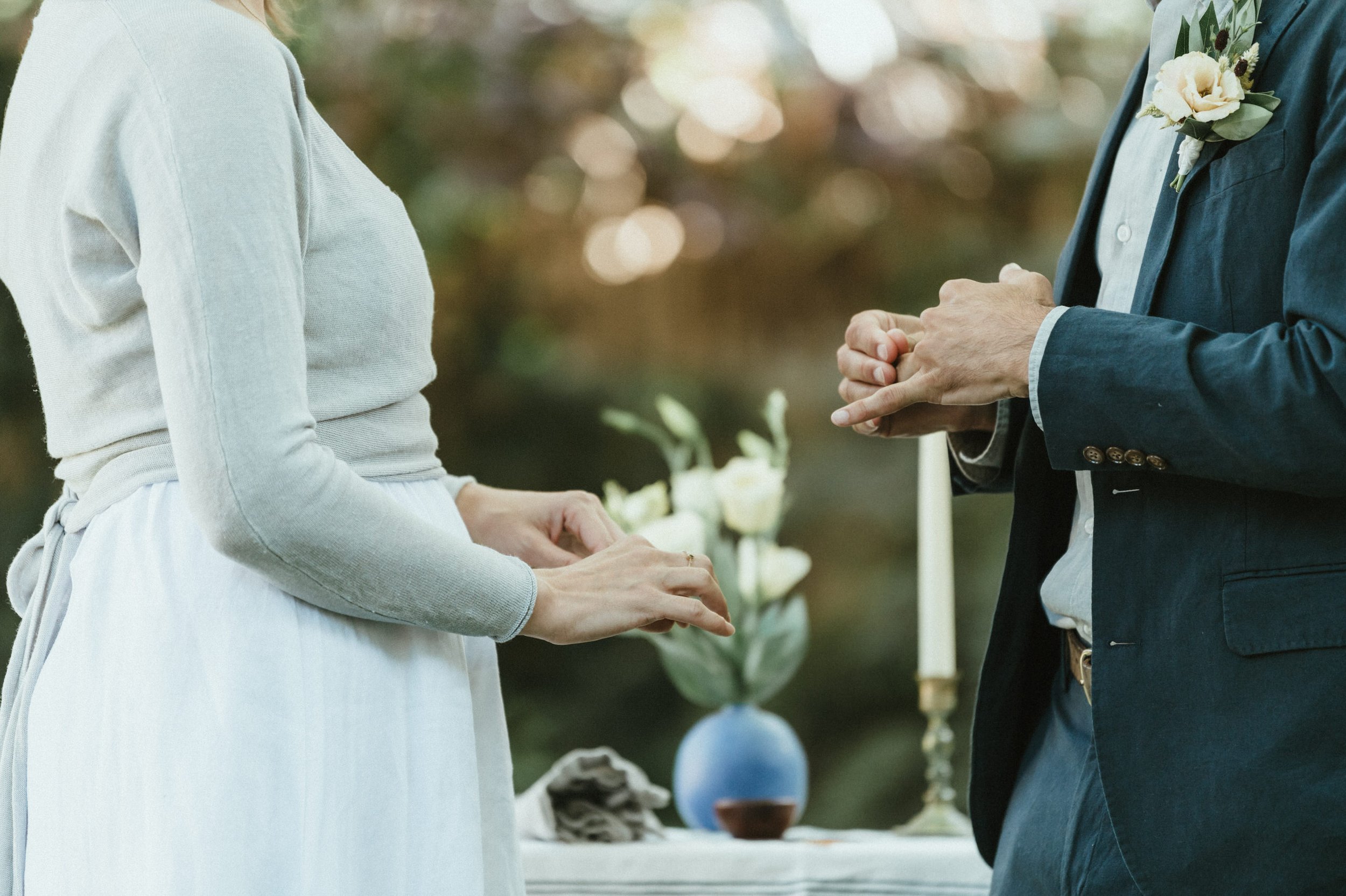 bride and groom hands with candle and flowers in the background exchanging rings